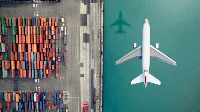 An overhead view of an airplane flying above cargo containers at a port